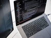 A laptop displaying lines of code on its screen, viewed from above. A second monitor is partially visible to the left, also showing code. Both devices are connected by cables on a light-colored desk. code sprint community typo3 code