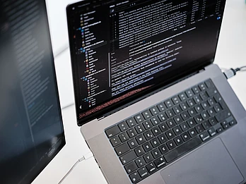 A laptop displaying lines of code on its screen, viewed from above. A second monitor is partially visible to the left, also showing code. Both devices are connected by cables on a light-colored desk. code sprint community typo3 code