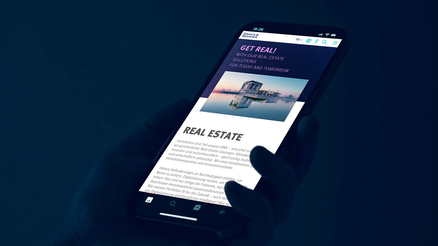 A hand holding a smartphone displaying the Drees & Sommer mobile website showing the market real estate titled “GET REAL!”. Featuring an image of a modern building and text about their real estate services, emphasizing innovative and sustainable solutions. The background is dark, creating a focus on the illuminated screen.